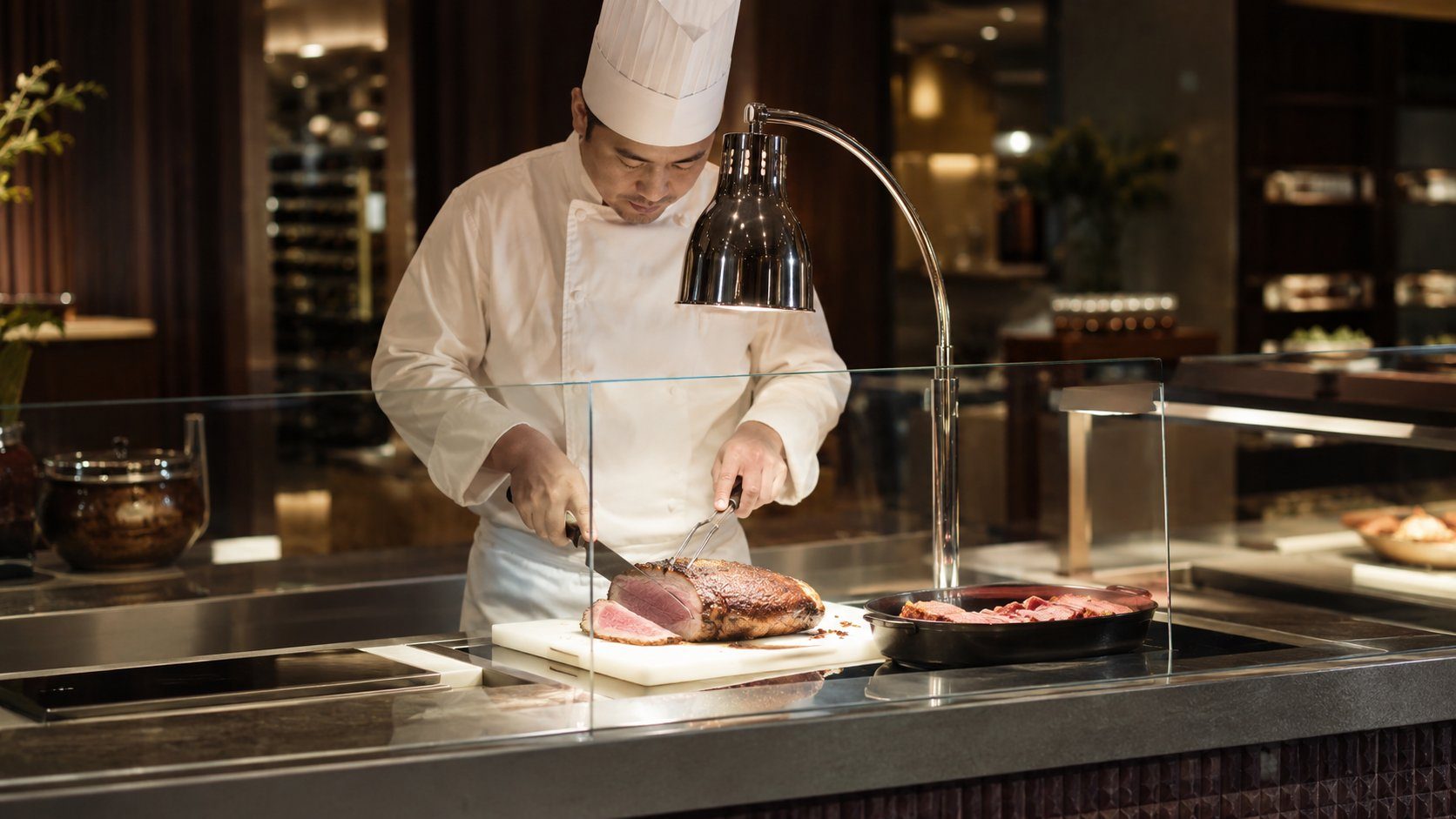 Chef carving a roast at a live-cooking station on a hotel buffet line under a heat lamp, behind a glass sneeze guard